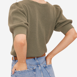 A woman wearing a ribbed short-sleeve knit top in olive green, paired with light blue jeans, shown from a side-back angle against a plain light background.