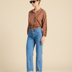 A woman modeling a brown and orange patterned button-up shirt with blue cropped jeans and dark brown flat shoes, standing against a solid beige studio background.