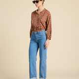A woman modeling a brown and orange patterned button-up shirt with blue cropped jeans and dark brown flat shoes, standing against a solid beige studio background.