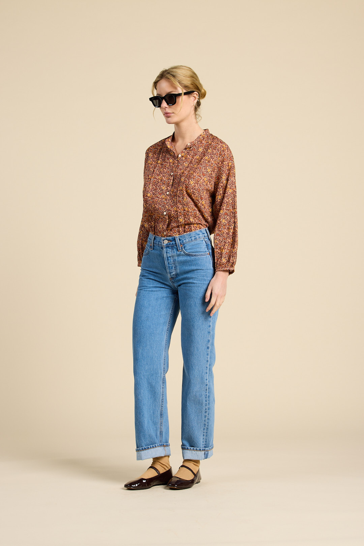 A woman modeling a brown and orange patterned button-up shirt with blue cropped jeans and dark brown flat shoes, standing against a solid beige studio background.