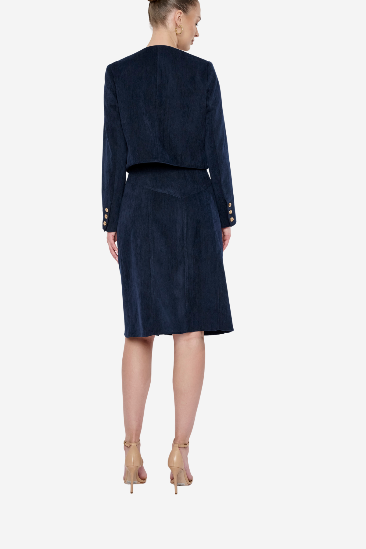A model shown from the back wearing a navy blue velvet two-piece set consisting of a cropped jacket with gold button details on the sleeves and a matching knee-length skirt, paired with beige high-heeled sandals against a light gray background.