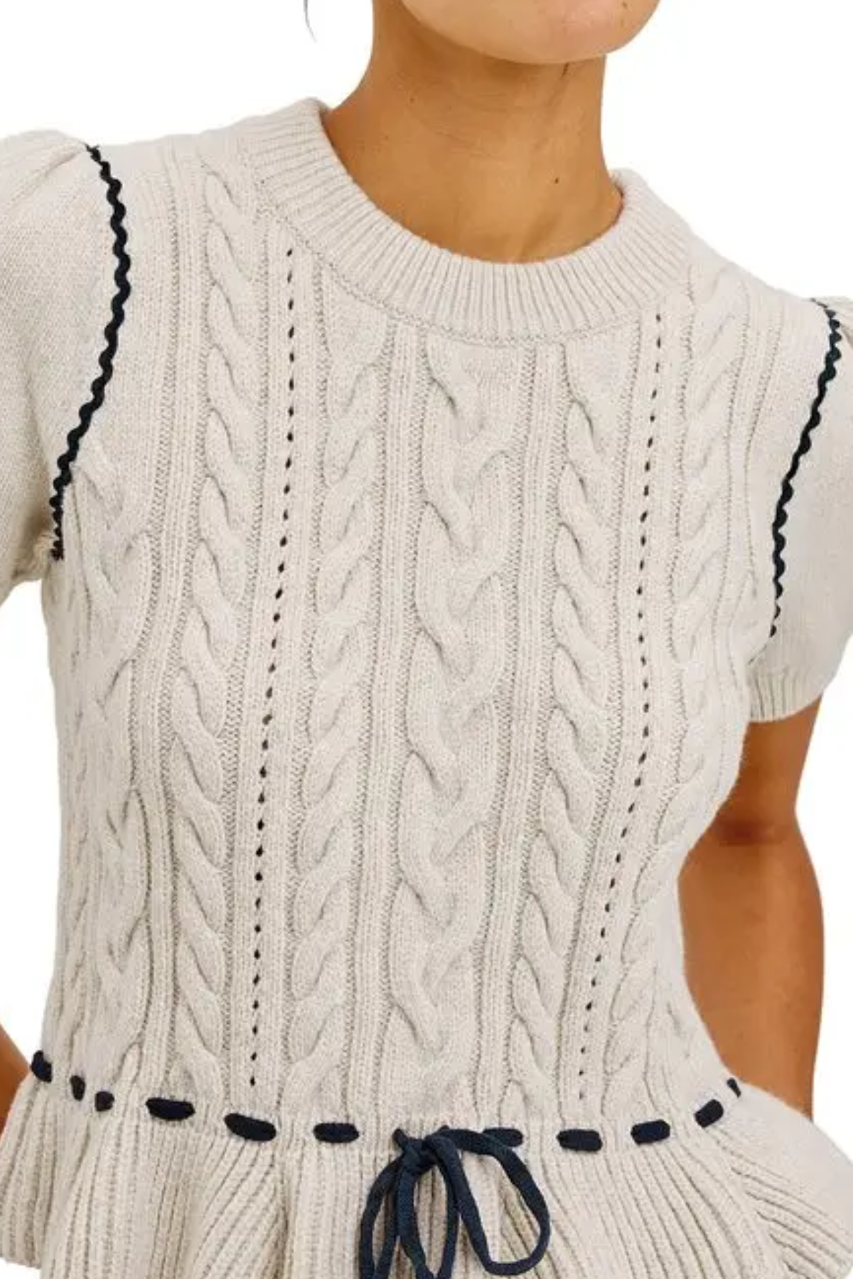 Close-up of a cream cable-knit short-sleeve top or dress with dark navy wavy trim along the shoulders and waist, worn by a model.