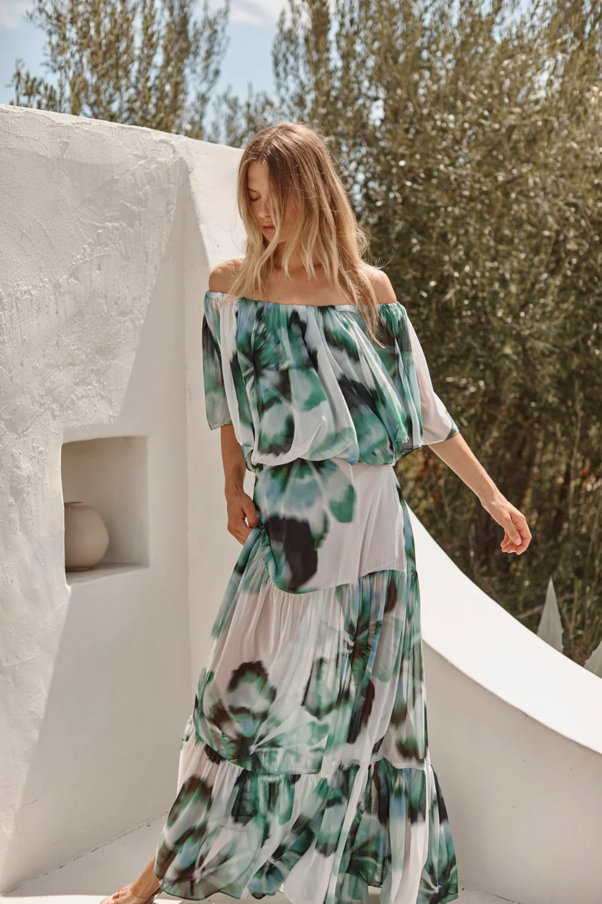 Woman wearing a floral dress standing against a white wall with greenery in the background