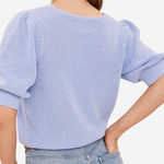 A woman wearing a light blue ribbed knit sweater with puffed elbow-length sleeves, shown from the back against a plain light background.