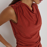 A close-up of a woman wearing a sleeveless rust-red top with a draped cowl neckline, gathered shoulder details, and ruching at the waist, paired with black bottoms against a light gray background.
