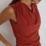 A close-up of a woman wearing a sleeveless rust-red top with a draped cowl neckline, gathered shoulder details, and ruching at the waist, paired with black bottoms against a light gray background.