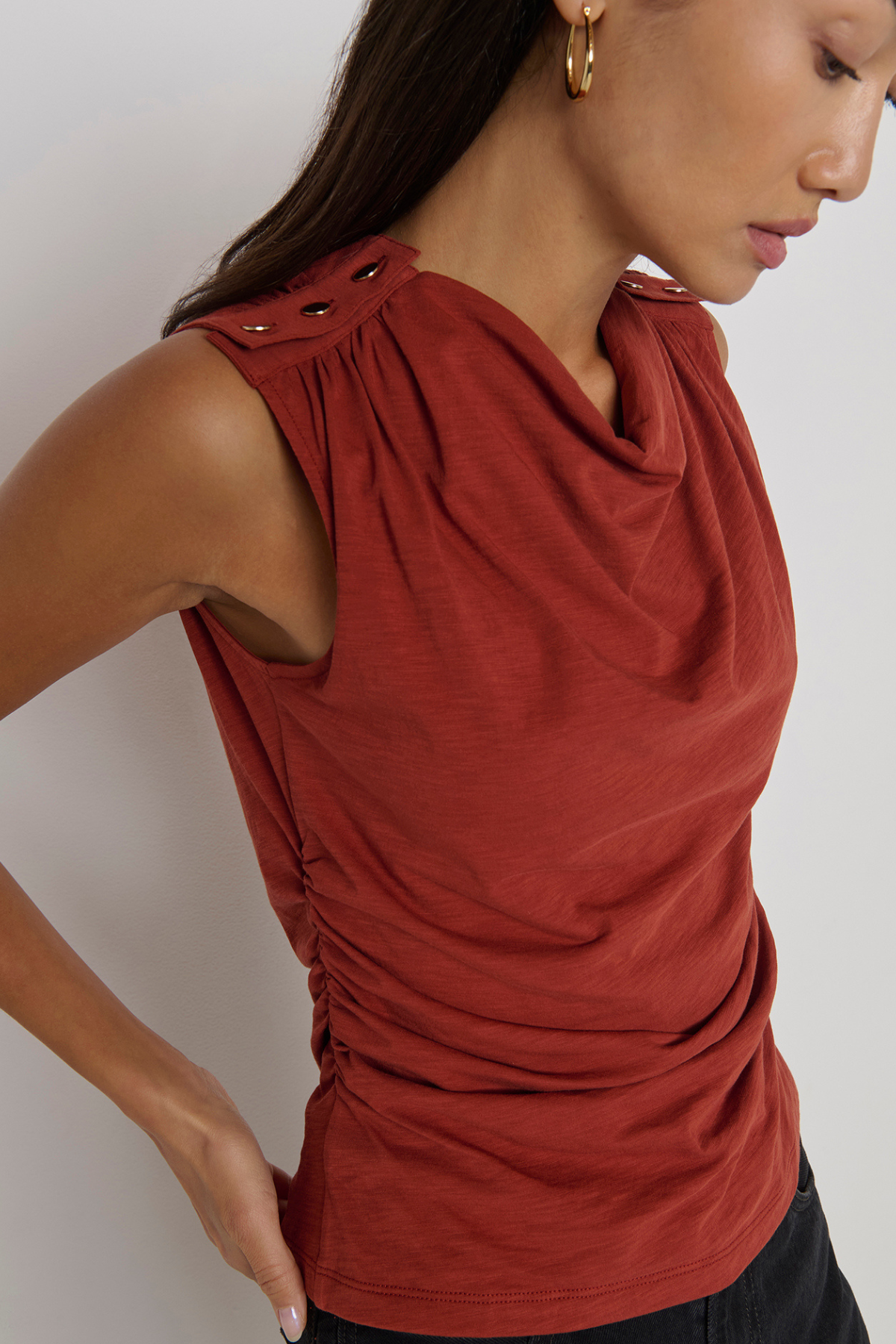 A close-up of a woman wearing a sleeveless rust-red top with a draped cowl neckline, gathered shoulder details, and ruching at the waist, paired with black bottoms against a light gray background.