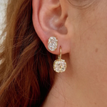 Close-up of a person's ear wearing a pair of gold hoop earrings with cushion-cut clear gemstones.