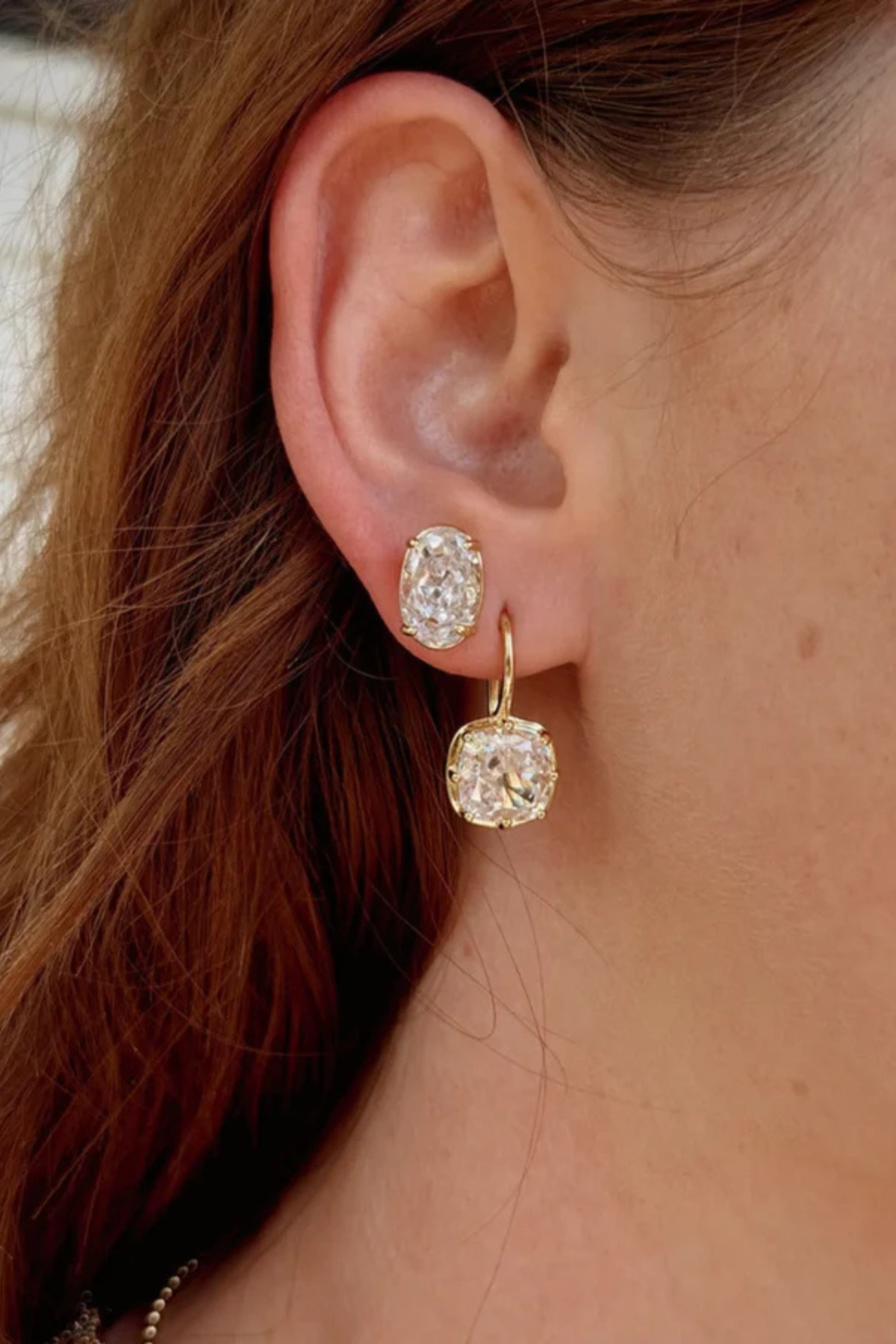 Close-up of a person's ear wearing a pair of gold hoop earrings with cushion-cut clear gemstones.