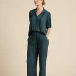 A woman modeling a matching dark green two-piece lounge set with a small white geometric pattern, consisting of a short-sleeve button-up top and wide-leg trousers, set against a solid beige studio background.
