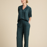 A woman modeling a matching dark green two-piece lounge set with a small white geometric pattern, consisting of a short-sleeve button-up top and wide-leg trousers, set against a solid beige studio background.