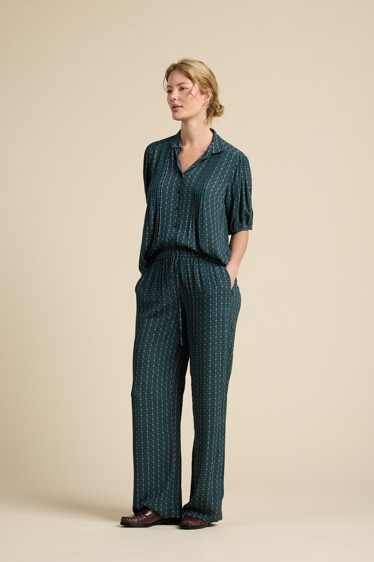 A woman modeling a matching dark green two-piece lounge set with a small white geometric pattern, consisting of a short-sleeve button-up top and wide-leg trousers, set against a solid beige studio background.