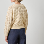 Woman wearing a beige cable knit sweater and dark blue jeans on a plain background