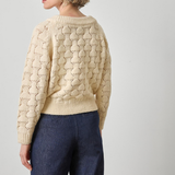 Woman wearing a beige cable knit sweater and dark blue jeans on a plain background