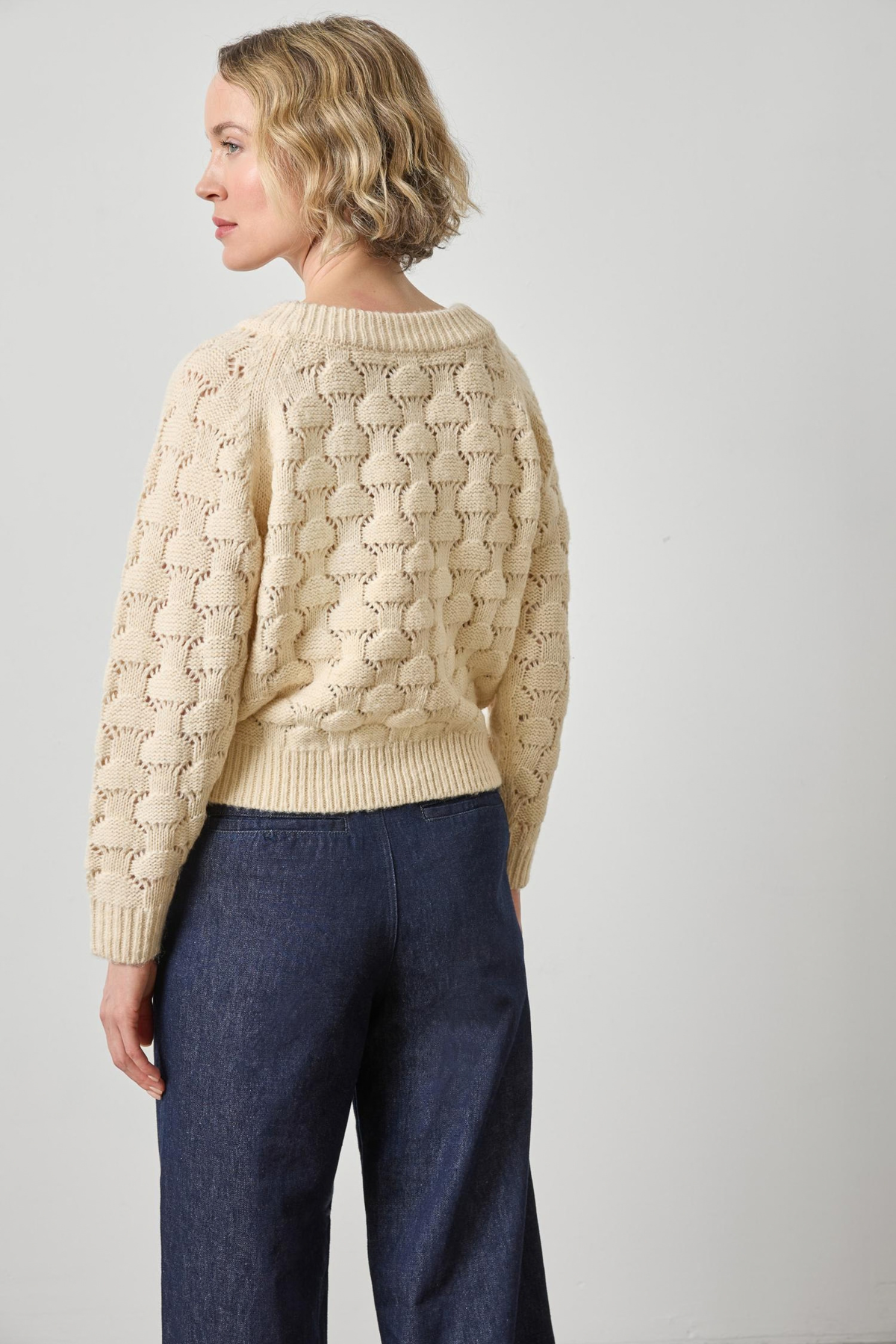 Woman wearing a beige cable knit sweater and dark blue jeans on a plain background