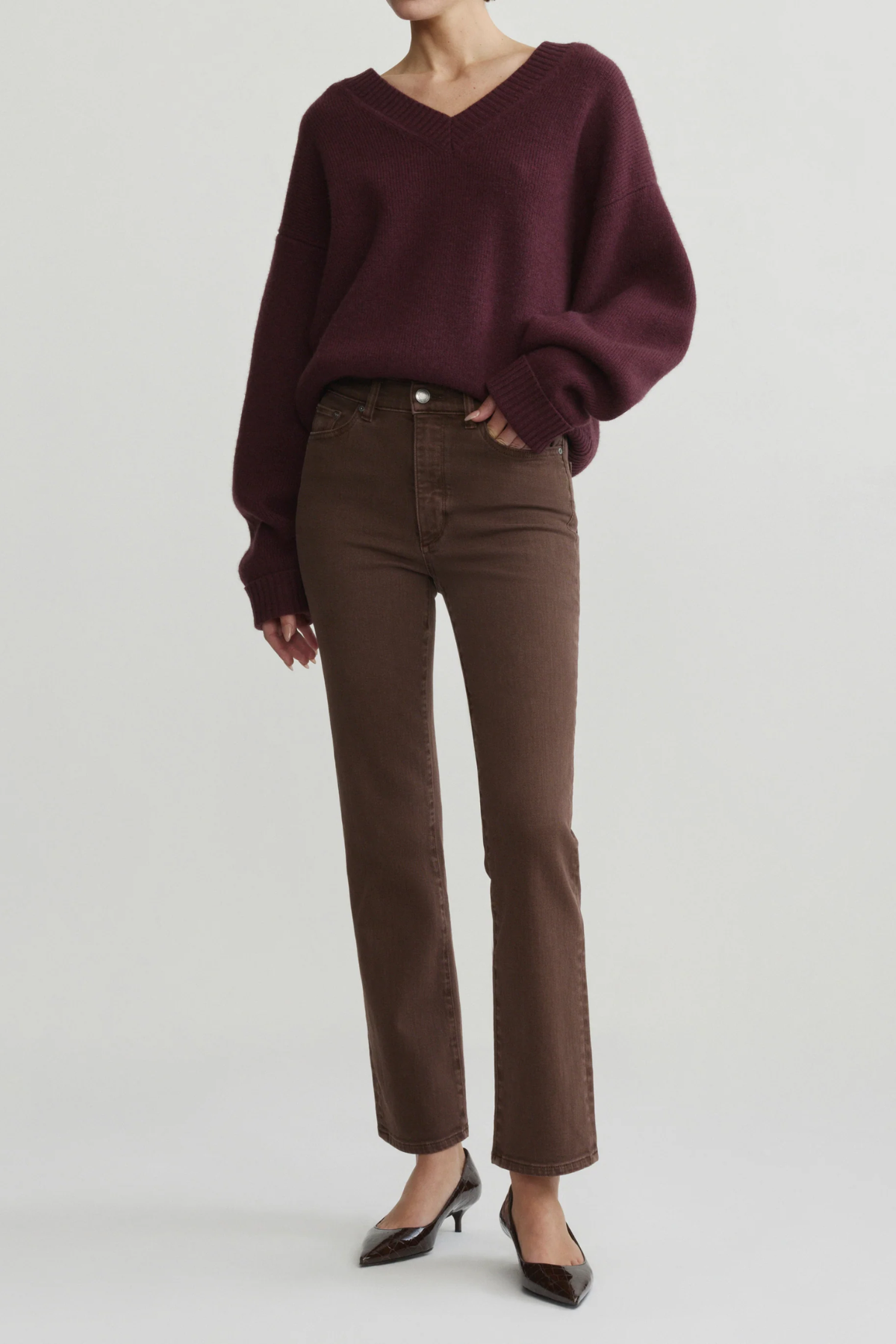 A model wearing a dark burgundy knit V-neck sweater and brown straight-leg jeans, paired with black pointed-toe heels, against a light gray studio background.