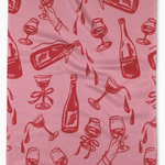 A pink waffle-weave kitchen towel featuring a repeating red pattern of champagne bottles, wine glasses, and hands holding glasses.