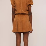 A model shown from the back wearing a matching tan-brown short-sleeve top and pleated mini skirt set, photographed against a plain off-white studio background.