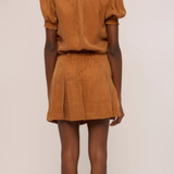 A model shown from the back wearing a matching tan-brown short-sleeve top and pleated mini skirt set, photographed against a plain off-white studio background.