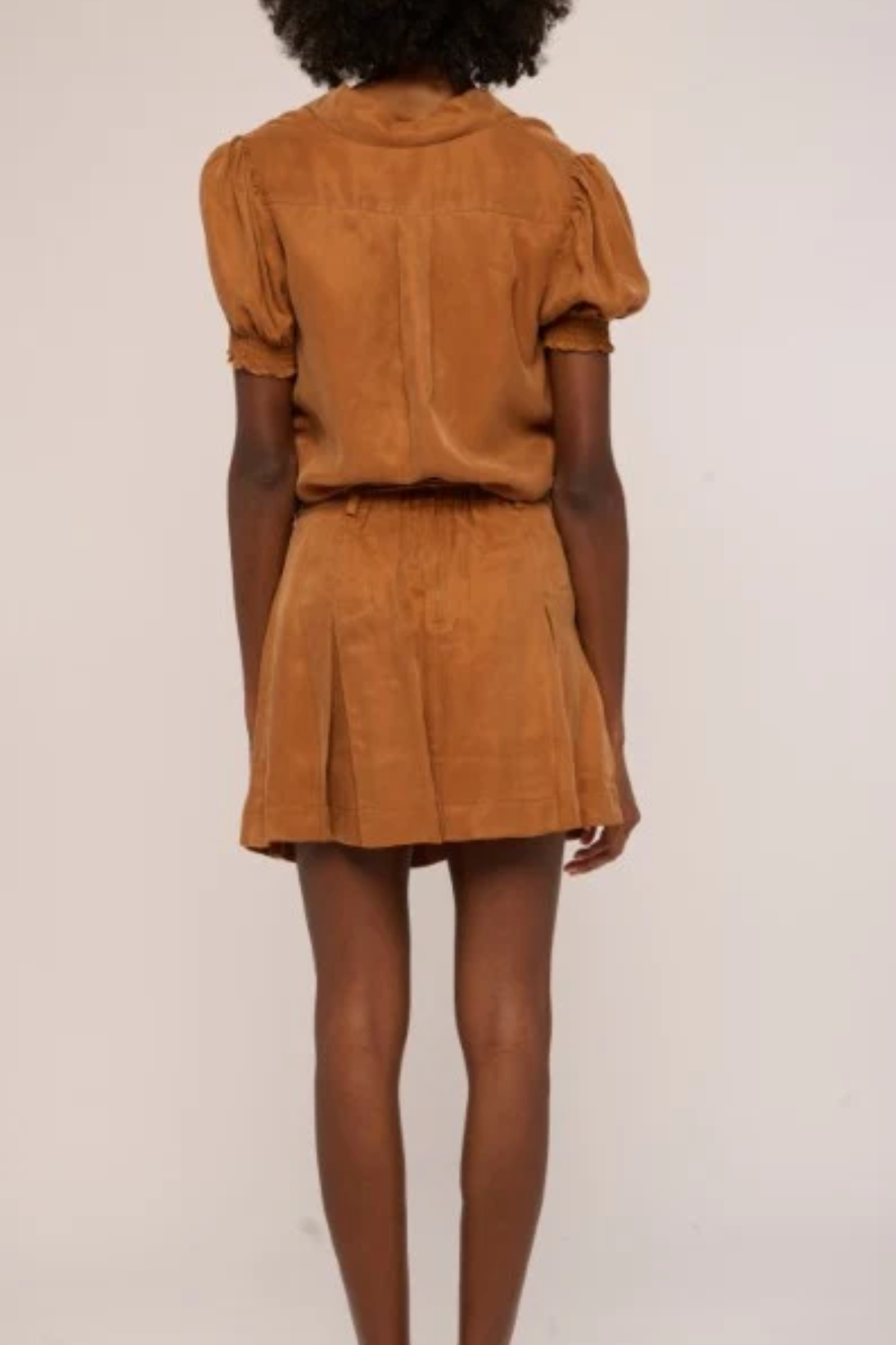 A model shown from the back wearing a matching tan-brown short-sleeve top and pleated mini skirt set, photographed against a plain off-white studio background.