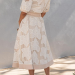 Woman wearing a beige lace dress against a white wall.
