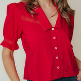 A woman wearing a bright red short-sleeve blouse with lace trim and white buttons, paired with dark blue jeans, against a plain light beige background.