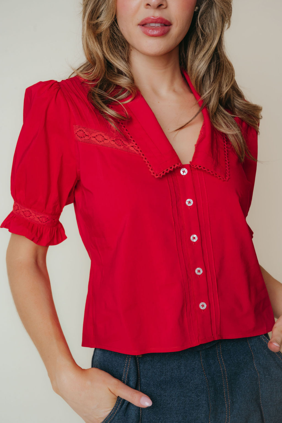 A woman wearing a bright red short-sleeve blouse with lace trim and white buttons, paired with dark blue jeans, against a plain light beige background.