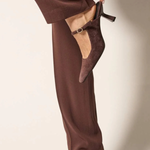 A person wearing dark brown wide-leg trousers and matching pointed-toe kitten heel slingback shoes, with one hand lifting the side of the trouser leg to show the shoe more clearly against a plain light beige studio background.