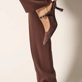 A person wearing dark brown wide-leg trousers and matching pointed-toe kitten heel slingback shoes, with one hand lifting the side of the trouser leg to show the shoe more clearly against a plain light beige studio background.