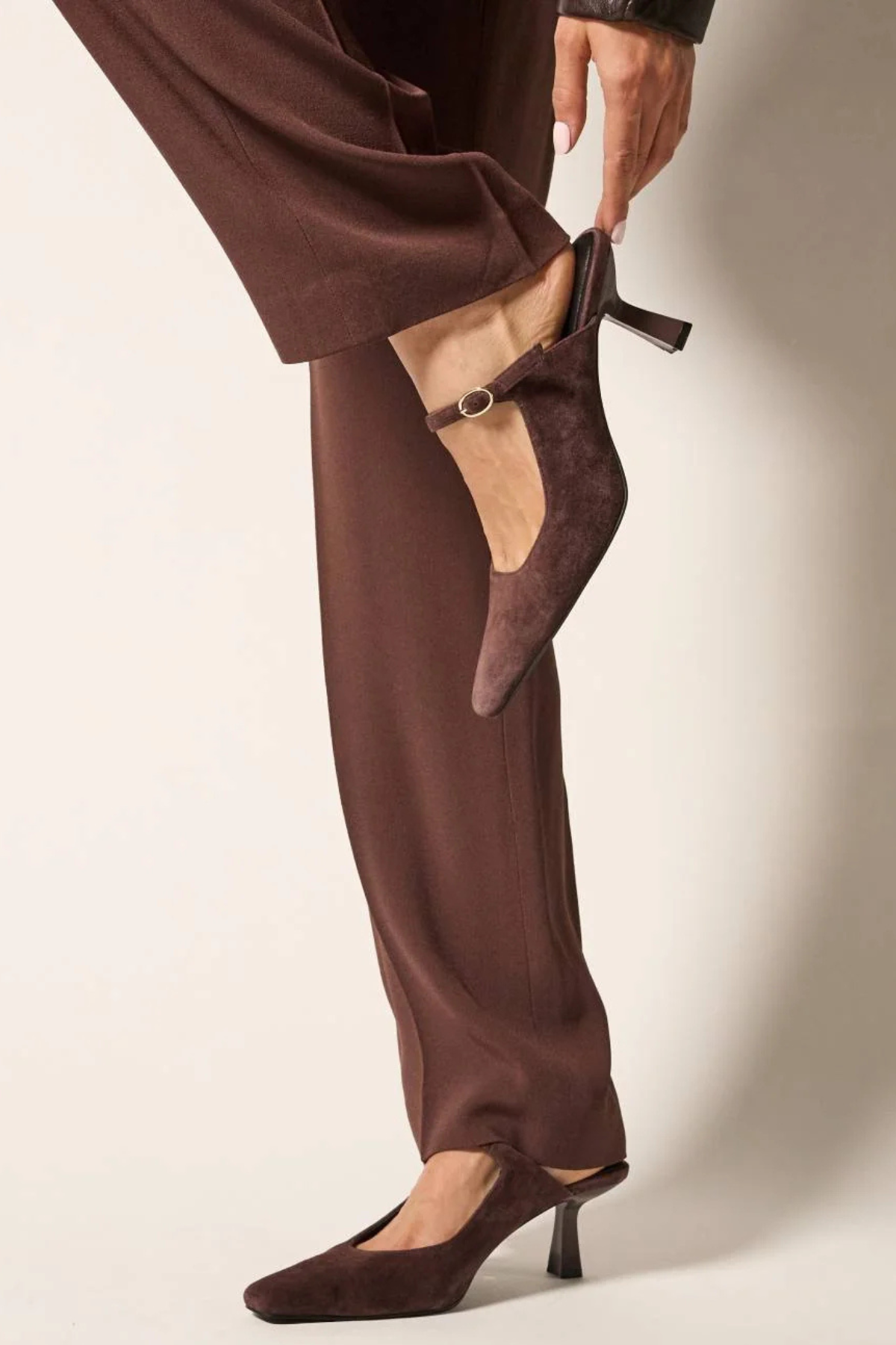 A person wearing dark brown wide-leg trousers and matching pointed-toe kitten heel slingback shoes, with one hand lifting the side of the trouser leg to show the shoe more clearly against a plain light beige studio background.