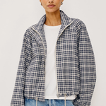 A woman modeling a cropped, long-sleeve plaid jacket with a high collar, worn over a white t-shirt and blue jeans against a light gray studio background.