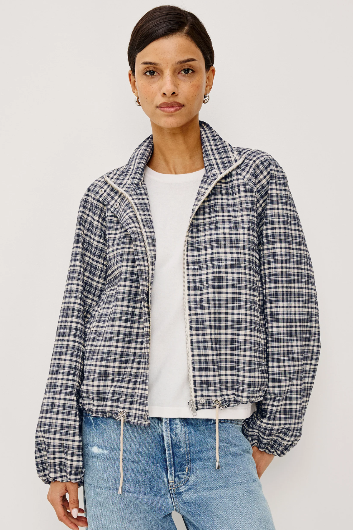 A woman modeling a cropped, long-sleeve plaid jacket with a high collar, worn over a white t-shirt and blue jeans against a light gray studio background.