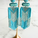 A pair of long beaded fringe earrings in turquoise and light blue, displayed on a gold stand against a white marble background.