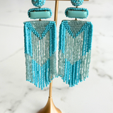 A pair of long beaded fringe earrings in turquoise and light blue, displayed on a gold stand against a white marble background.