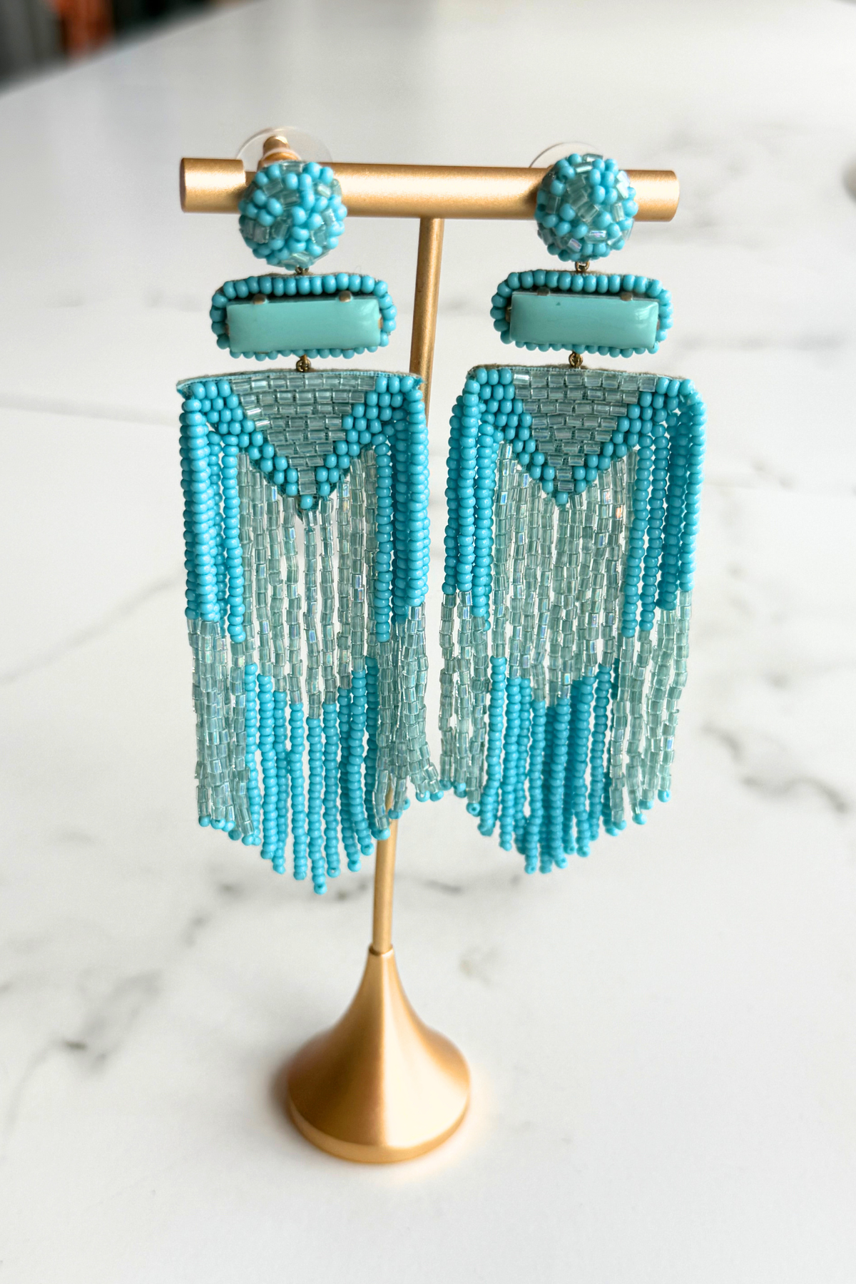 A pair of long beaded fringe earrings in turquoise and light blue, displayed on a gold stand against a white marble background.