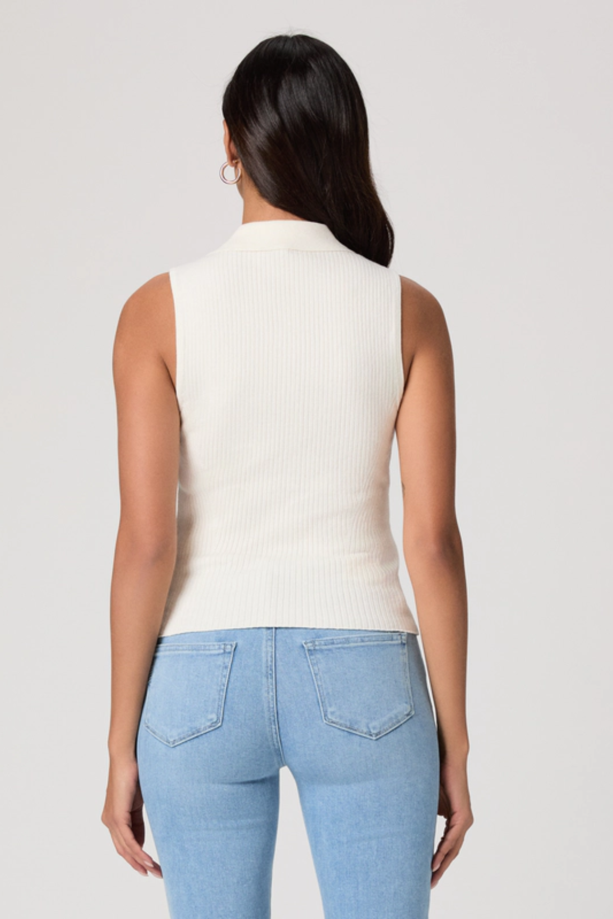 Woman wearing a white sleeveless top and light blue jeans on a plain background