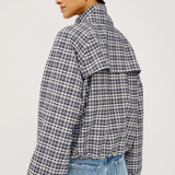 A woman shown from the back wearing a navy and white plaid cropped jacket with voluminous sleeves, paired with light blue jeans, against a plain light gray studio background.