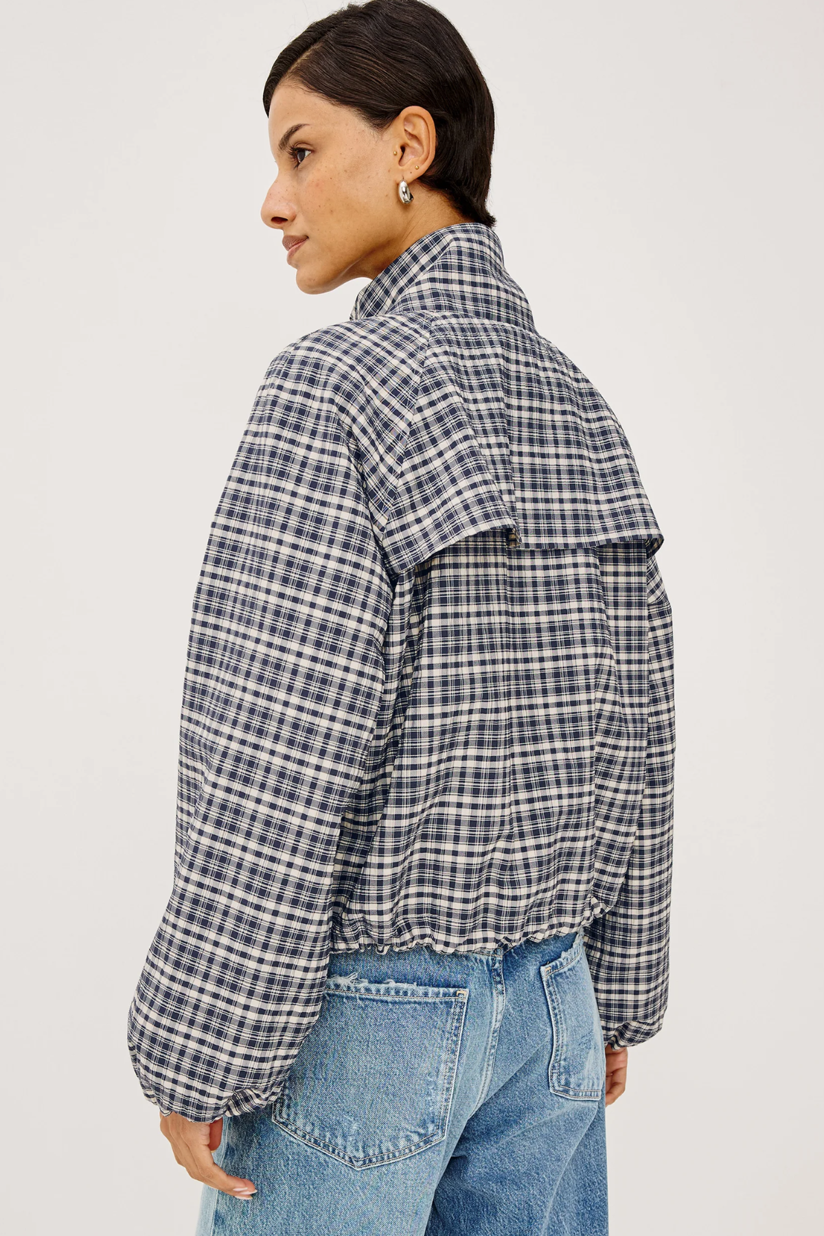 A woman shown from the back wearing a navy and white plaid cropped jacket with voluminous sleeves, paired with light blue jeans, against a plain light gray studio background.