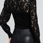 A model shown from the back wearing a black long-sleeve lace blouse with structured shoulders and black faux-leather pants, against a light gray studio background.