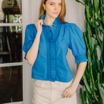 Woman wearing a blue blouse and beige pants standing next to a cactus plant indoors.
