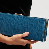 A person in a black velvet dress holds a blue textured clutch bag with gold-tone hardware against a light gray background.