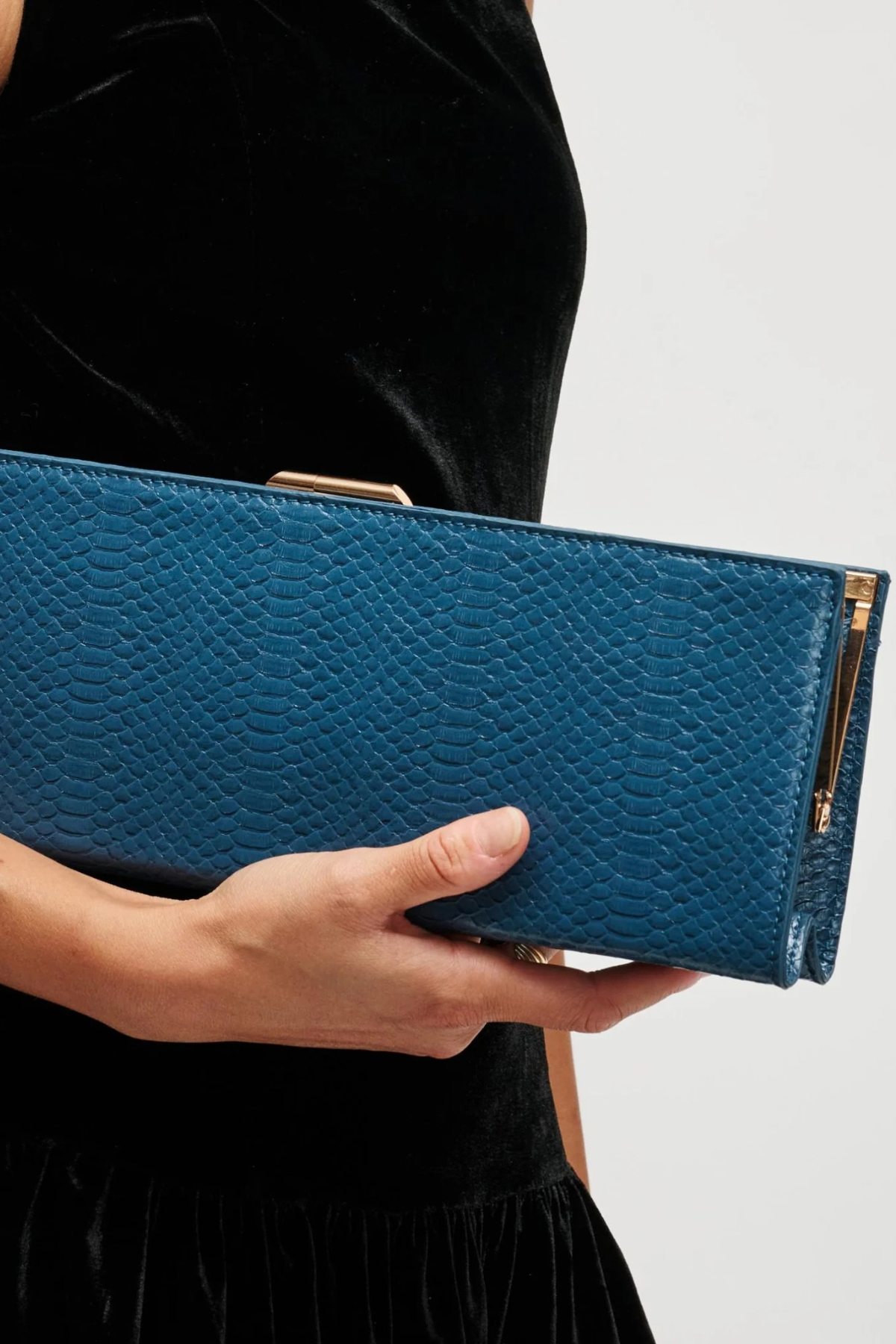 A person in a black velvet dress holds a blue textured clutch bag with gold-tone hardware against a light gray background.