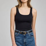 A woman modeling a black scoop-neck tank top paired with blue jeans and a black belt, photographed against a neutral light gray studio background.