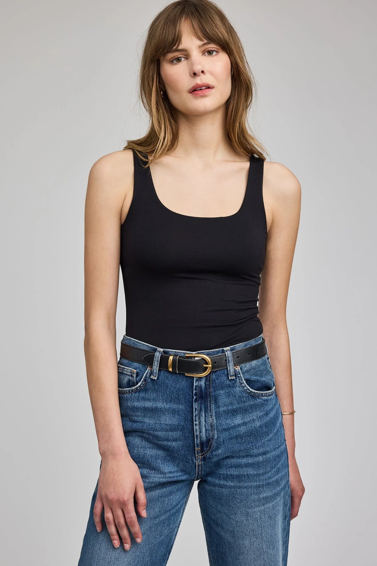 A woman modeling a black scoop-neck tank top paired with blue jeans and a black belt, photographed against a neutral light gray studio background.