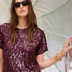 A woman wearing a sparkling burgundy sequined short-sleeve top and blue jeans, holding a large silver umbrella. She is wearing black sunglasses and a gold chain bracelet.