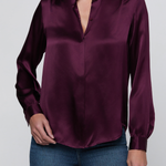 A woman wearing a long-sleeve satin blouse in deep burgundy with a split collar and button-front placket, paired with blue jeans, against a light gray studio background.