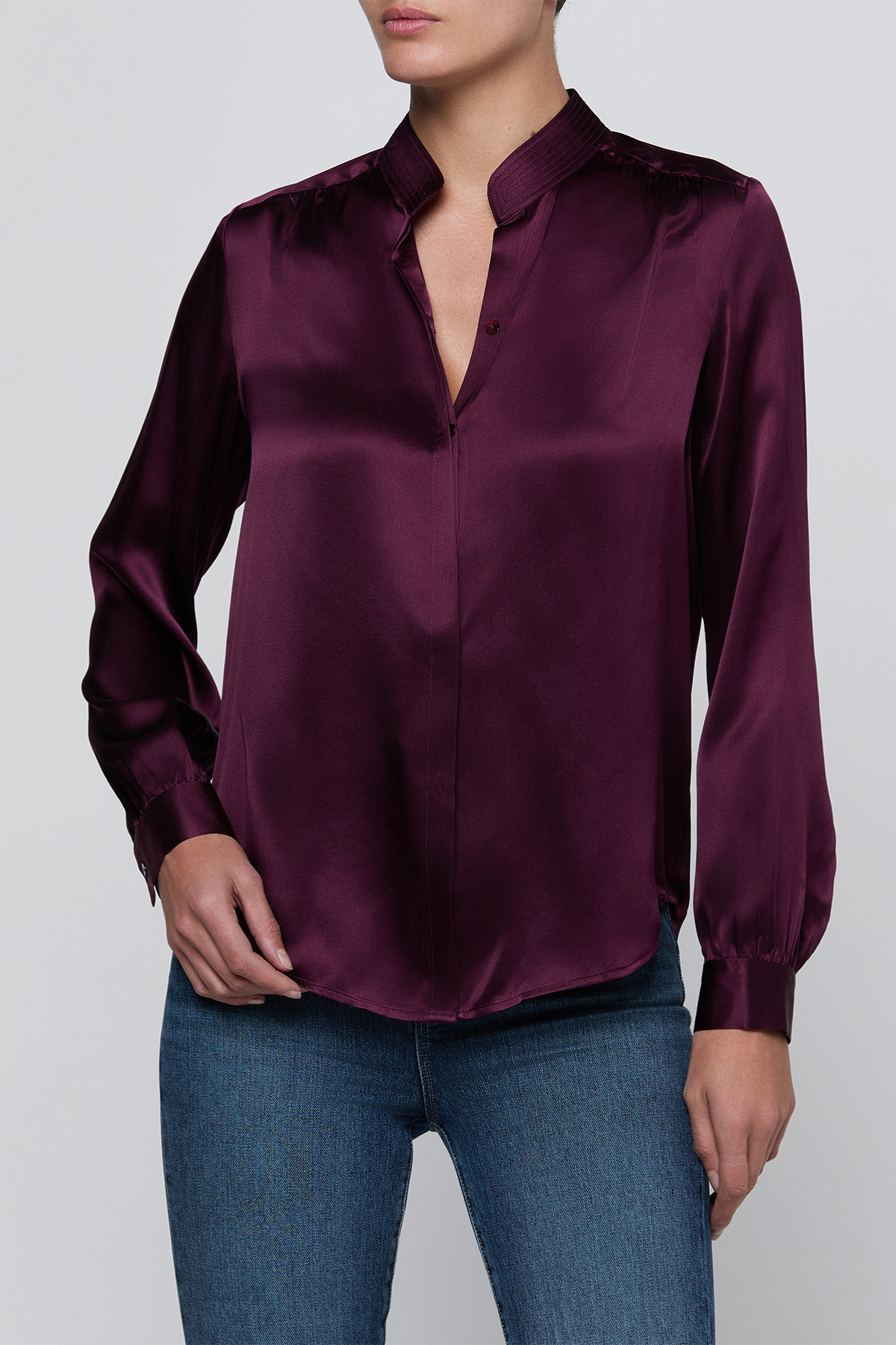 A woman wearing a long-sleeve satin blouse in deep burgundy with a split collar and button-front placket, paired with blue jeans, against a light gray studio background.
