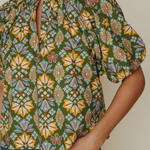 A close-up of a woman wearing a short-sleeve blouse with a green base and a colorful geometric floral pattern, paired with blue jeans.