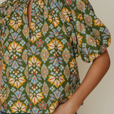 A close-up of a woman wearing a short-sleeve blouse with a green base and a colorful geometric floral pattern, paired with blue jeans.