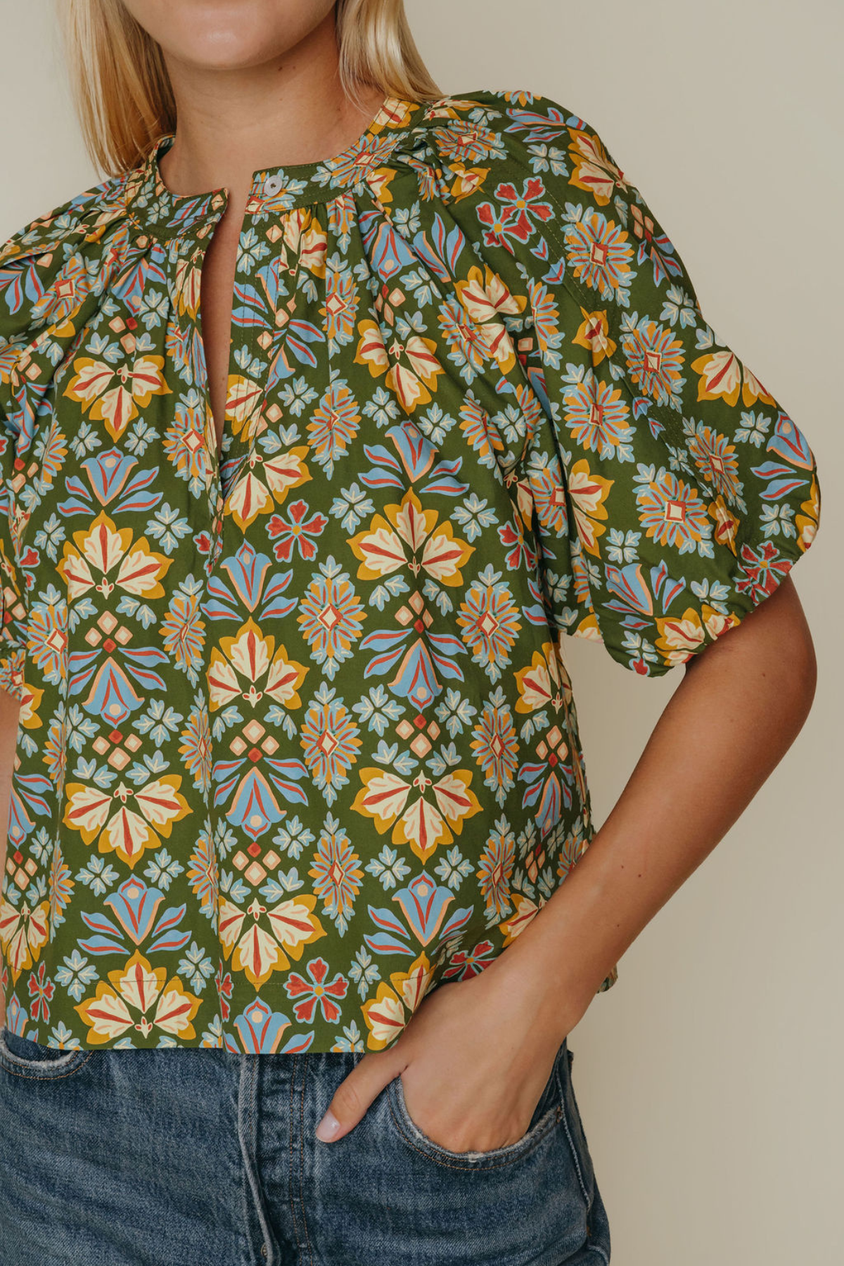 A close-up of a woman wearing a short-sleeve blouse with a green base and a colorful geometric floral pattern, paired with blue jeans.